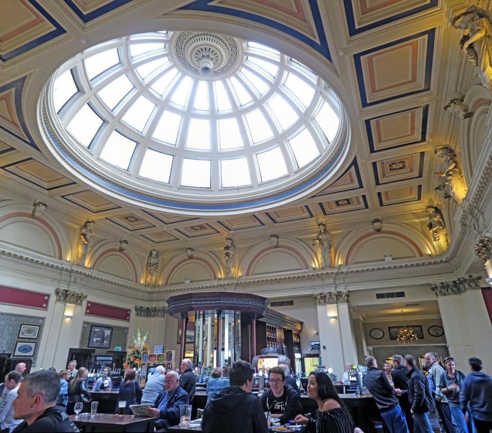 Interior of a pub with a large glass dome ceiling and patrons seated at the bar.