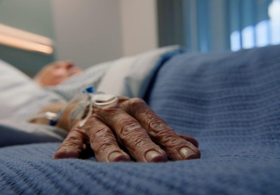 Close-up of an elderly man's hand resting on a hospital bed with an IV drip.