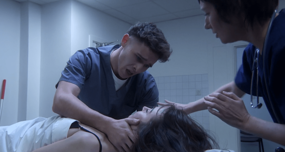 A distraught man and woman in scrubs attending to a patient lying on a bed.