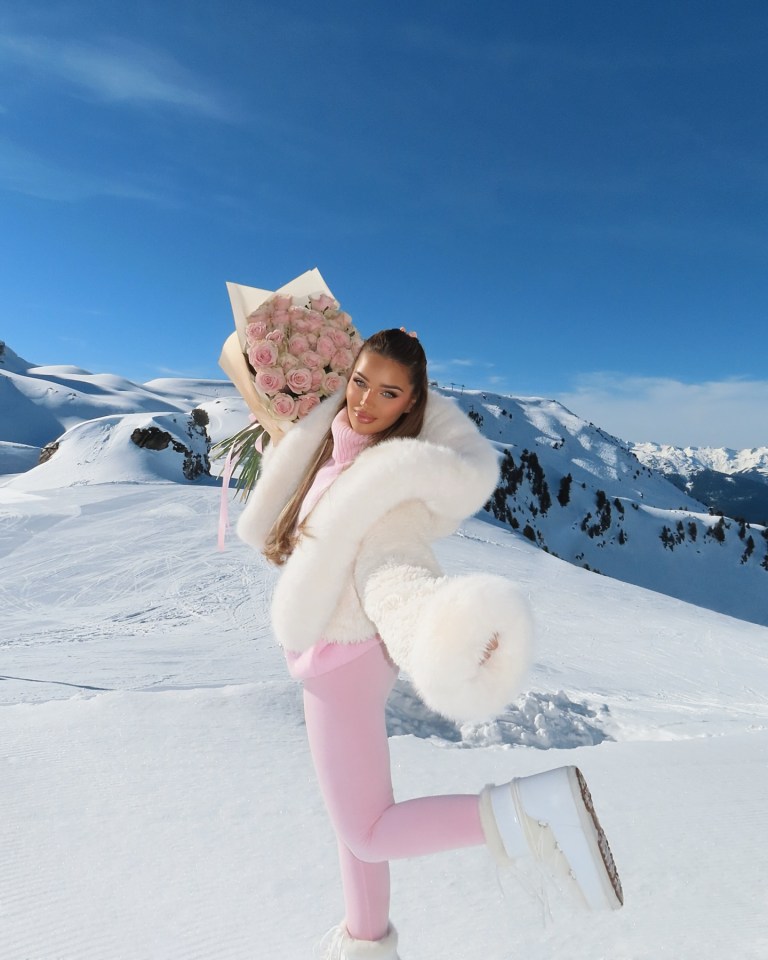 Woman in pink outfit and white fur jacket holding a large bouquet of roses in a snowy mountain landscape.