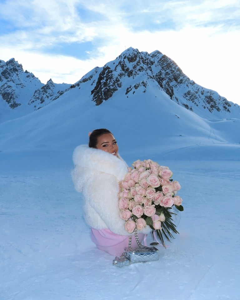 Woman in a white fur coat holding a large bouquet of pink roses in a snowy mountain landscape.