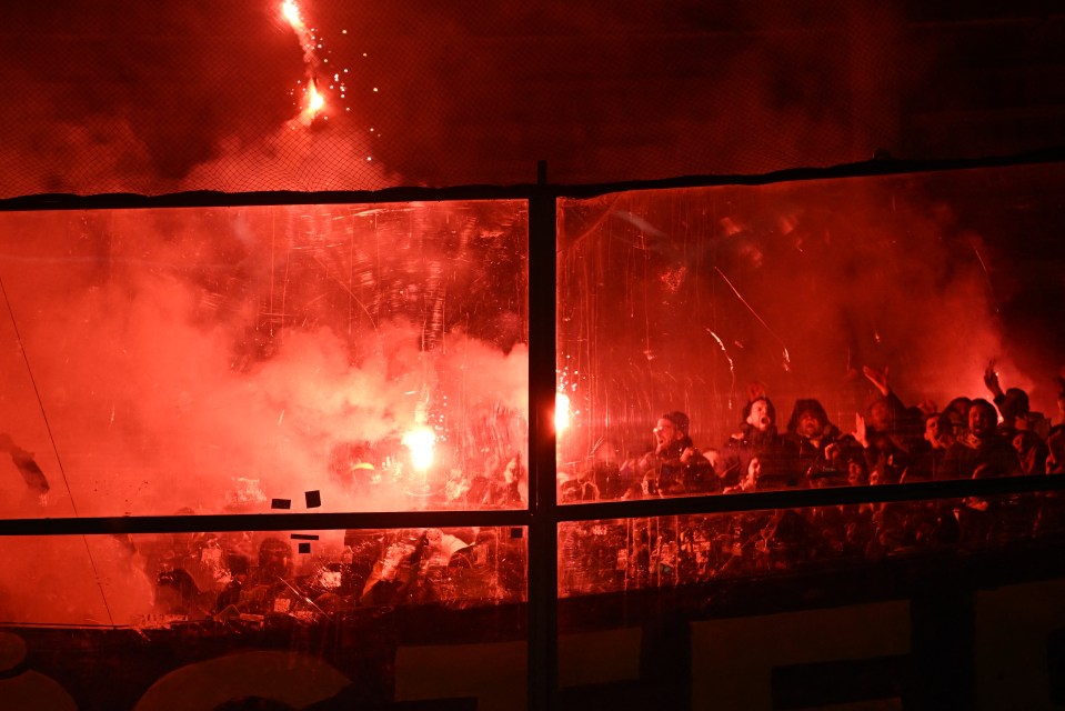 Fenerbahce supporters lighting red flares behind a protective barrier at a football match.