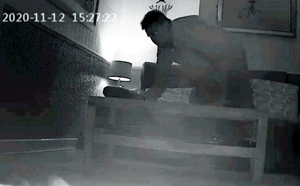 Black and white security camera footage of a man retrieving a recording device from a coffee table.