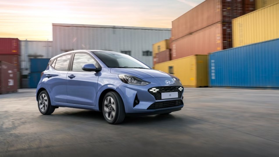A light blue Hyundai i10 driving past shipping containers.