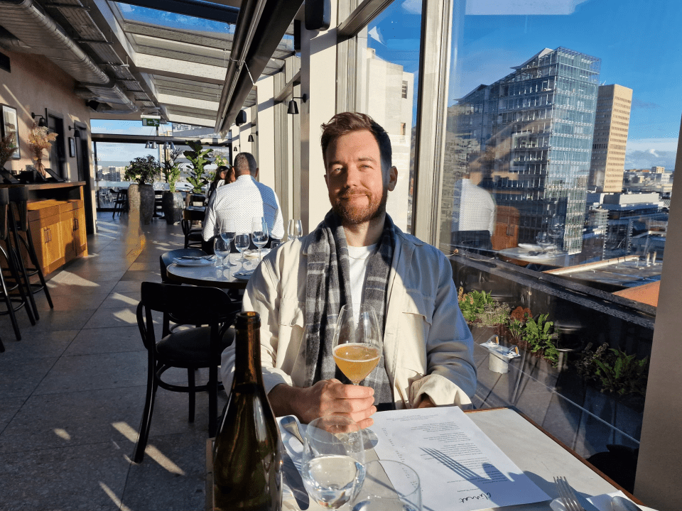 Man sitting at a restaurant table with a glass of wine, overlooking a city.