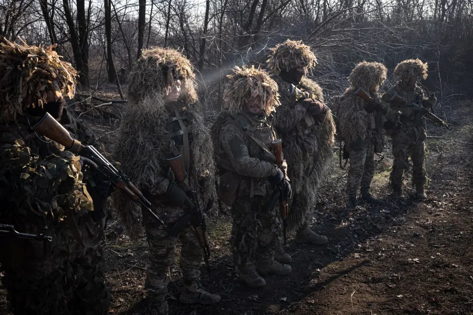 Soldiers in ghillie suits stand in a line holding rifles.