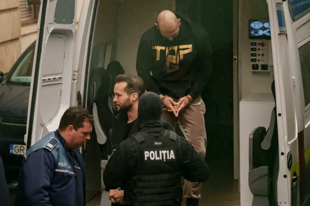 A handcuffed man in a black hoodie is escorted by police officers into a van.