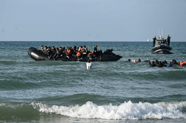 Migrants in a small boat and in the water, with a patrol boat in the background.