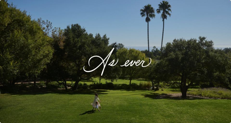 Woman and child walking on a grassy lawn with trees and palm trees in the background; text overlay: "As ever"