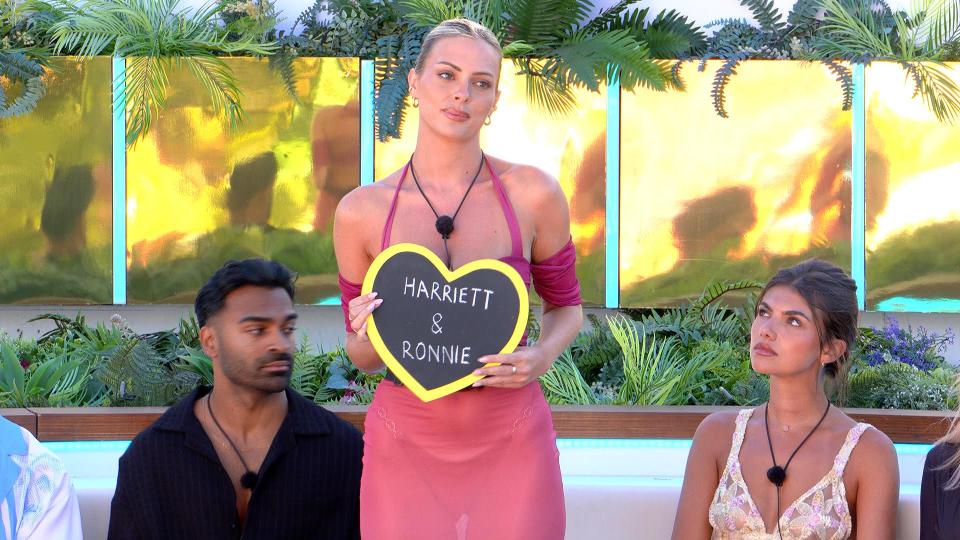 A woman on Love Island holds a sign that says "Harriett & Ronnie."