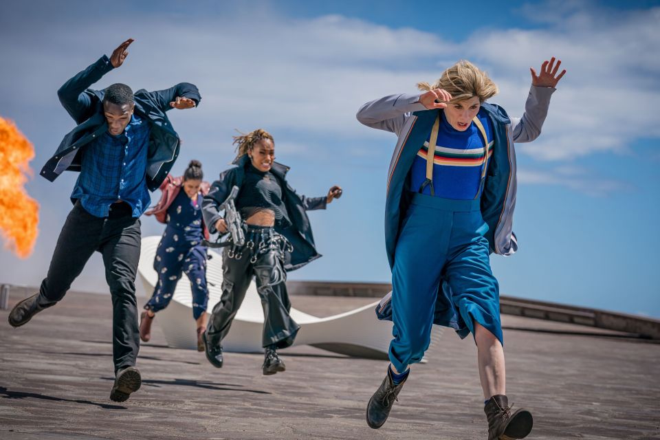 Jodie Whittaker as the Doctor running with companions from a fire.