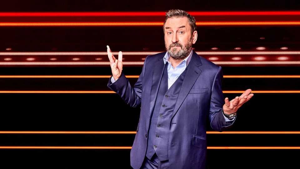 Lee Mack in front of The 1% Club logo.