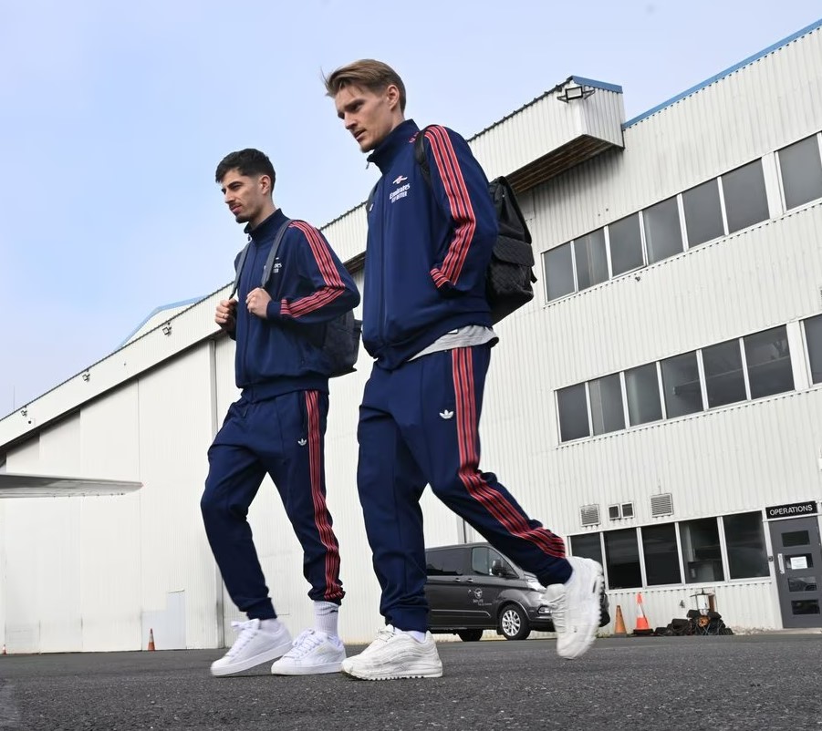 Two Arsenal players in tracksuits walking toward a vehicle.