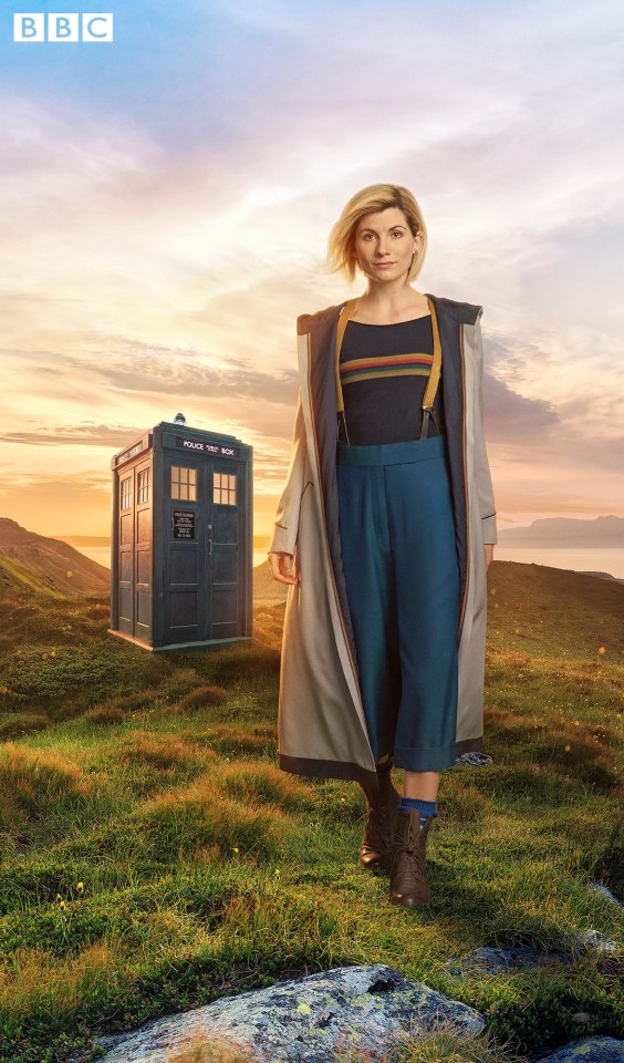 Jodie Whittaker as the Doctor in Doctor Who, standing in a field with the TARDIS in the background.
