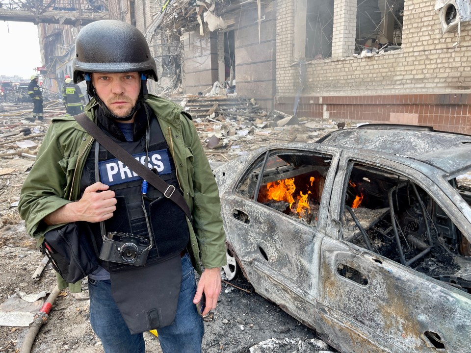 Journalist in protective gear at a fire in a war zone.