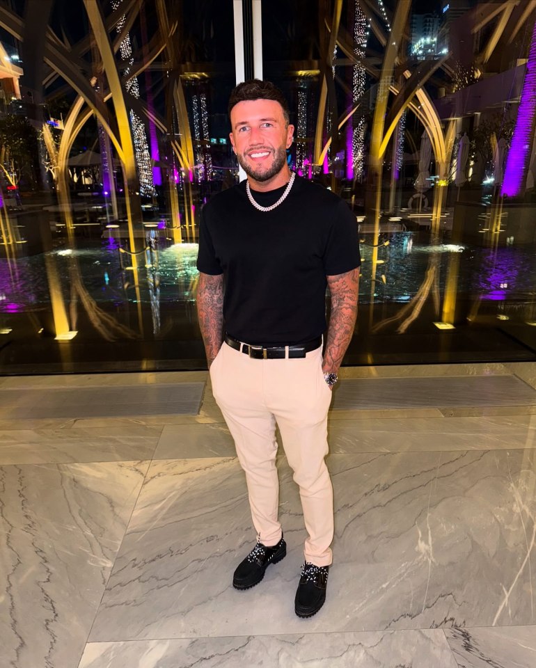A man in a black shirt and light beige pants stands in front of a large window at night.