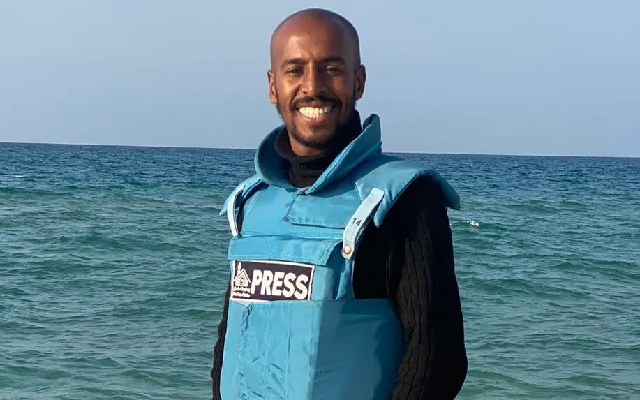 A cameraman wearing a blue press vest stands by the ocean.