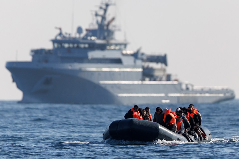 Migrants in a small boat crossing the English Channel, with a warship in the background.