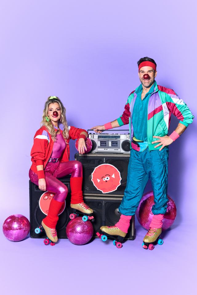 Two people in 80s-style roller skates and red noses pose with a boombox for Red Nose Day.