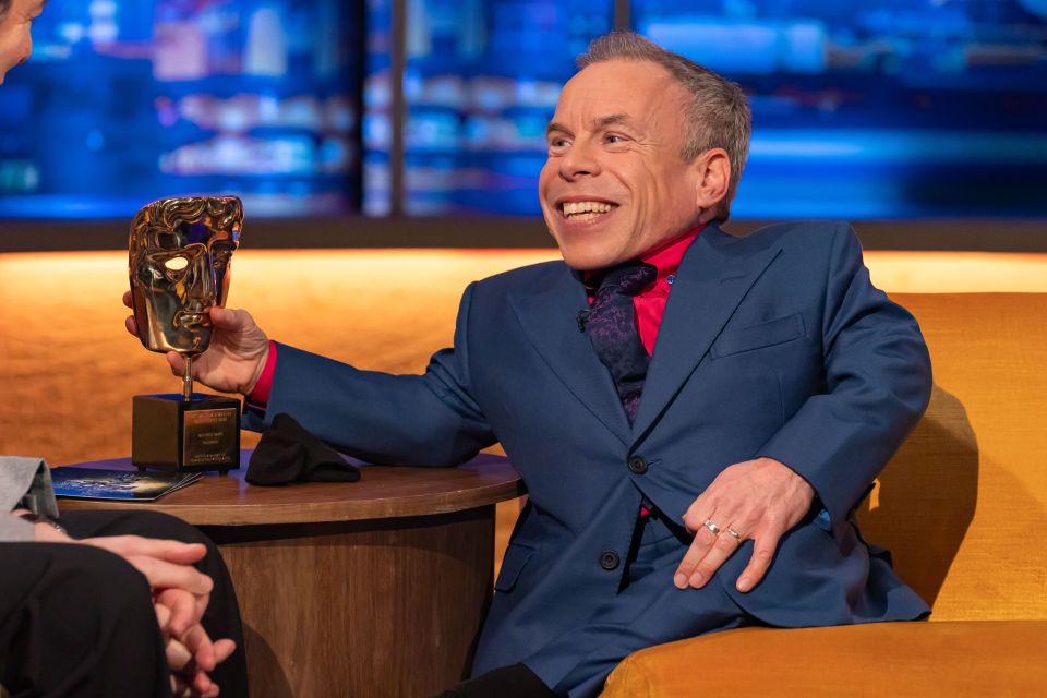 Warwick Davis holding a BAFTA award on a talk show.