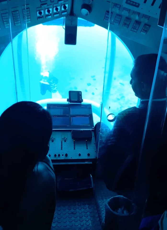 Interior view of a submarine, showing two passengers and a control panel, with an underwater scene visible through a large porthole.