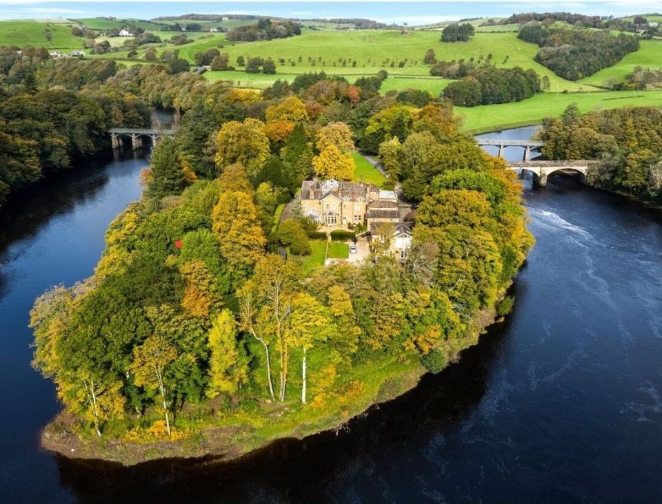 Aerial view of a house on a river island surrounded by autumn trees and two bridges.