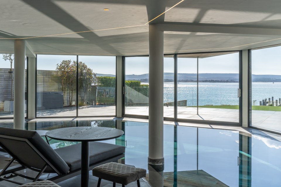 Indoor pool area with lounge chairs and a table overlooking a waterfront.