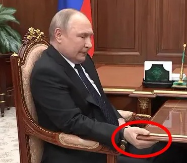 Vladimir Putin sitting at a large desk, his hand gripping the edge.