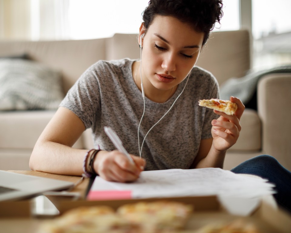Young woman studying at home while listening to music and eating pizza.