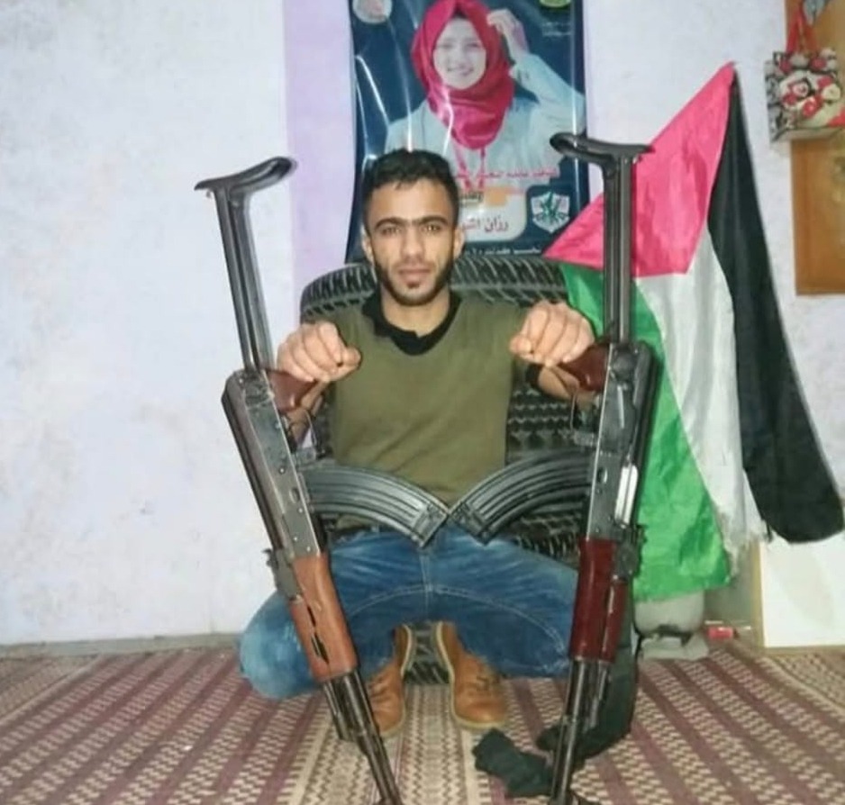 A man sits holding two rifles, a Palestinian flag behind him.