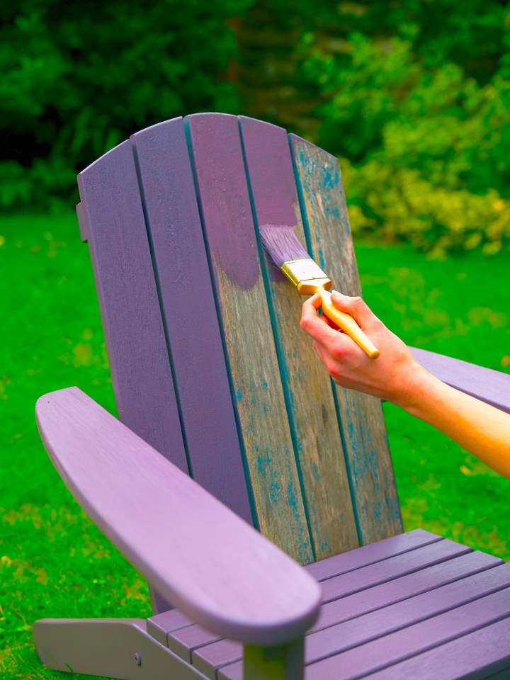 A person painting a purple Adirondack chair outdoors.