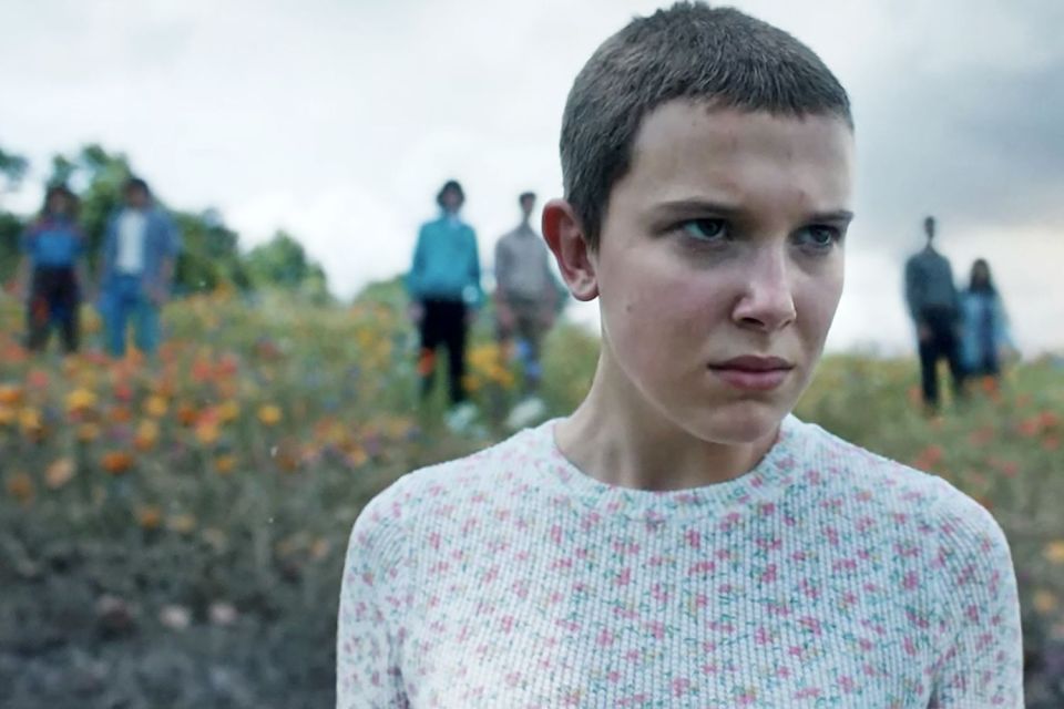Eleven from Stranger Things season 5, looking serious, in a field with other figures in the background.