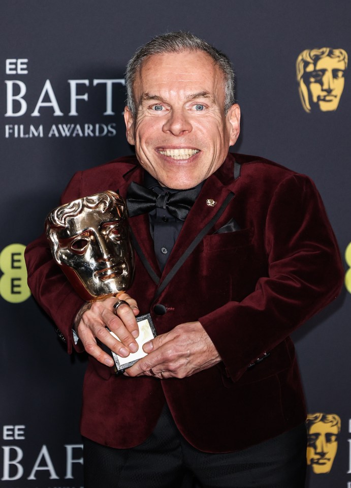 Warwick Davis holding a BAFTA award.