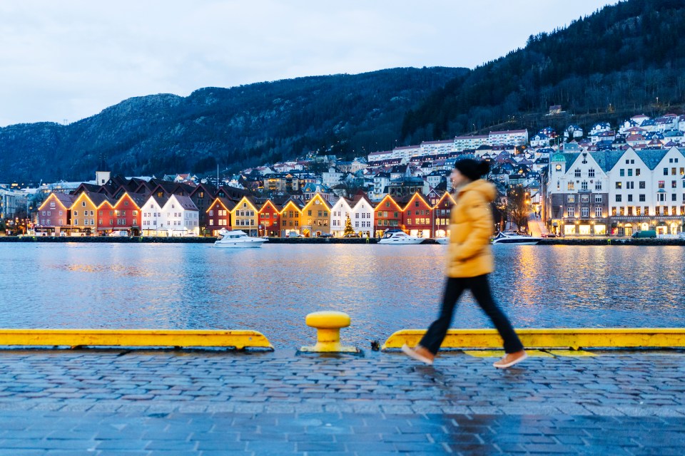 Person walking along a waterfront at dusk, past colorful buildings.