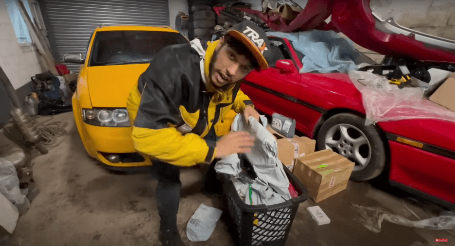 Person in yellow jacket unpacking a basket of car parts in a garage with a yellow car and a red car.