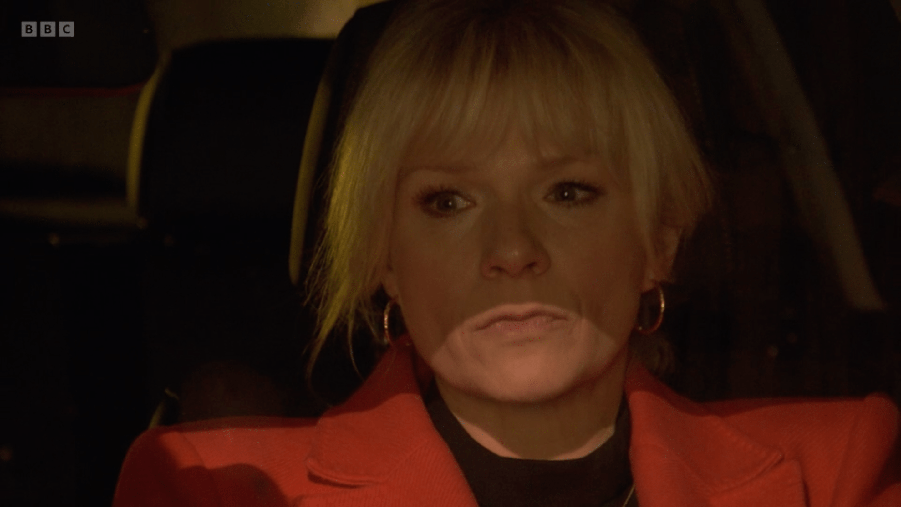 Screenshot of a blonde woman in a red jacket sitting in a car.