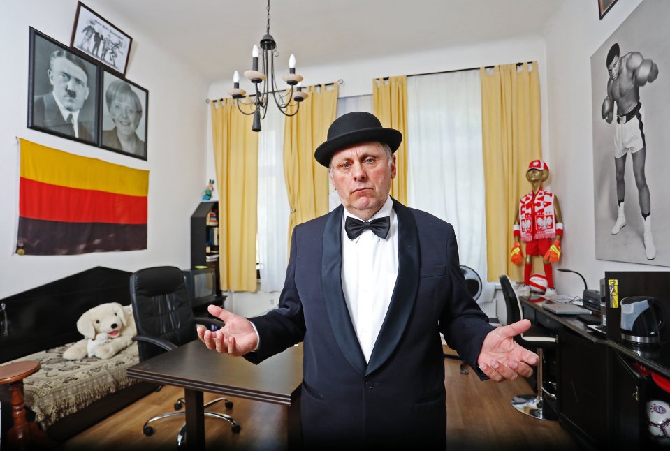 A man in a tuxedo and bowler hat stands with his arms outstretched in a room decorated with framed photos and a flag.