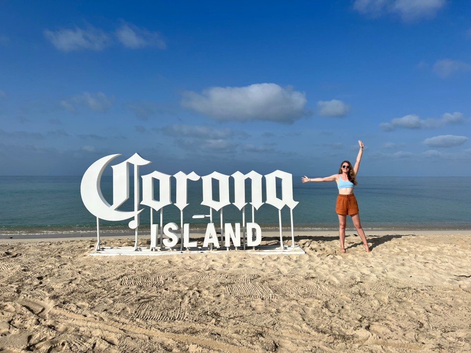 Woman standing in front of Corona Island sign on a beach.