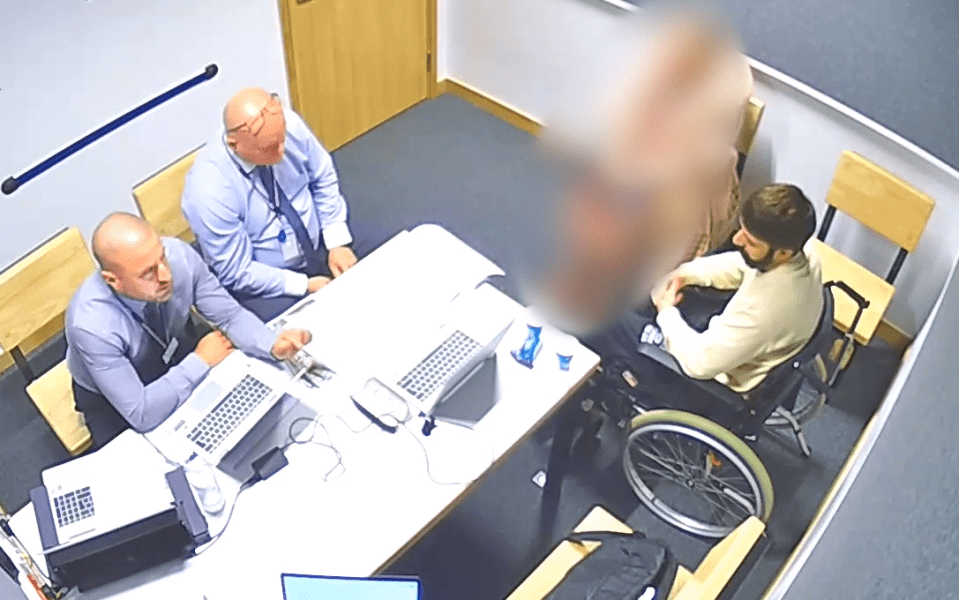 Overhead view of an interview; two men in suits sit at a table with laptops, facing a man in a wheelchair.