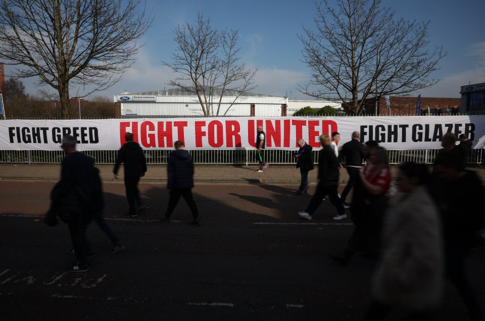 Person attaching a banner to a fence that reads "Fight Fight Fight For United Glazers Out" and depicts a skull and crossbones with a scarf.