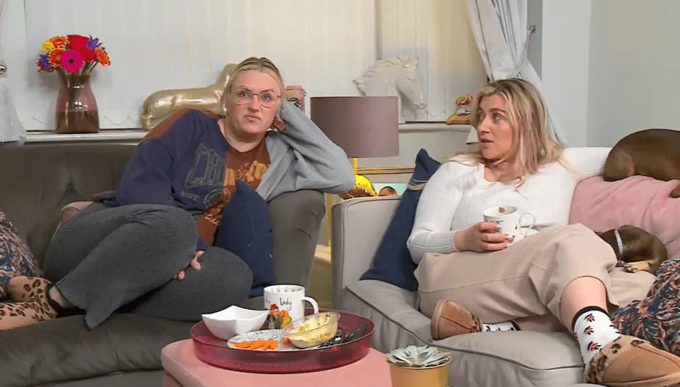 Two women sitting on a couch with dogs, drinking tea and eating snacks.