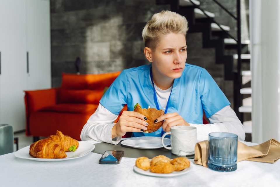 A female nurse in scrubs eats a hamburger and croissant during a break.