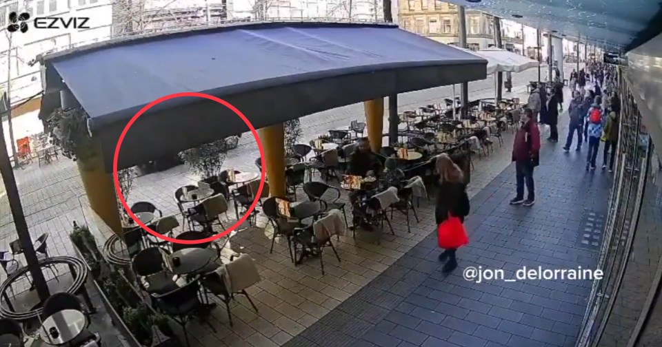 Security camera footage of a cafe patio with people sitting at tables and walking on the sidewalk.