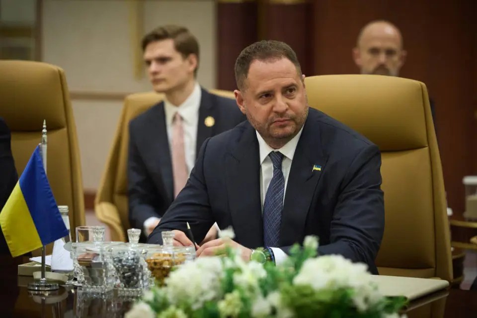 Man in suit sits at table, writing.  Ukrainian flag visible.