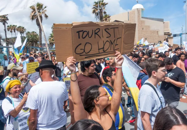 Protestor holding a sign that says "Tourist Go Home" at a demonstration.