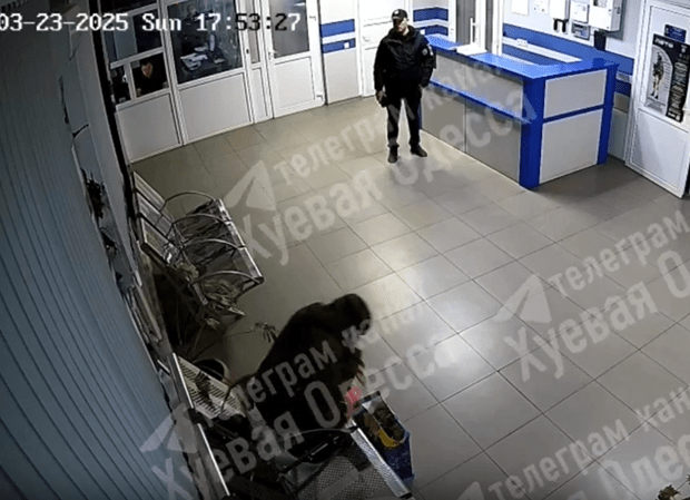 Security camera footage of a person bending over near a counter while a police officer stands nearby.
