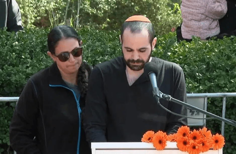 A man and woman stand at a podium with orange flowers in front of them; the man is speaking into a microphone.