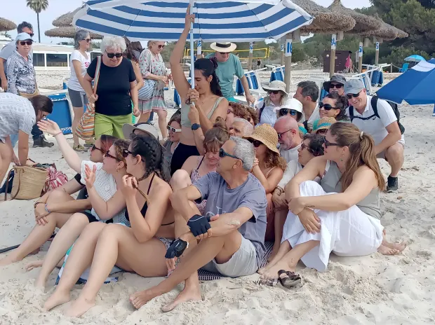 Large group of people sitting on a beach under a striped umbrella.