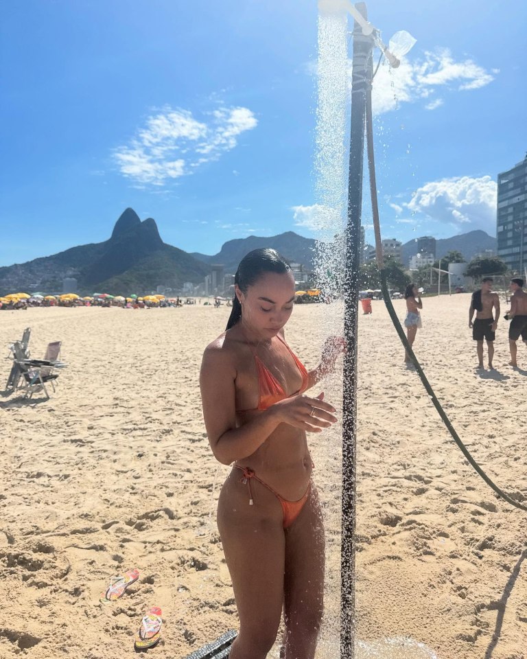 Woman in orange bikini rinsing off under an outdoor shower on a beach.
