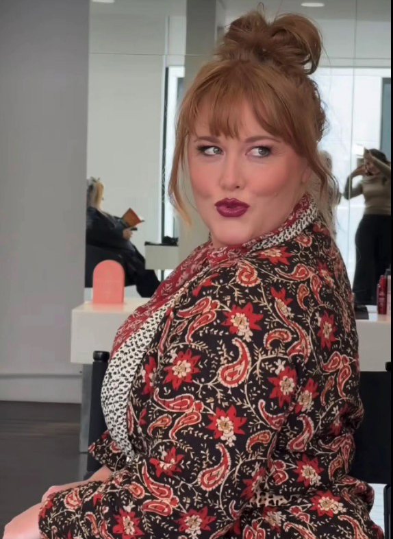Woman with red hair in a patterned shirt looking over her shoulder.